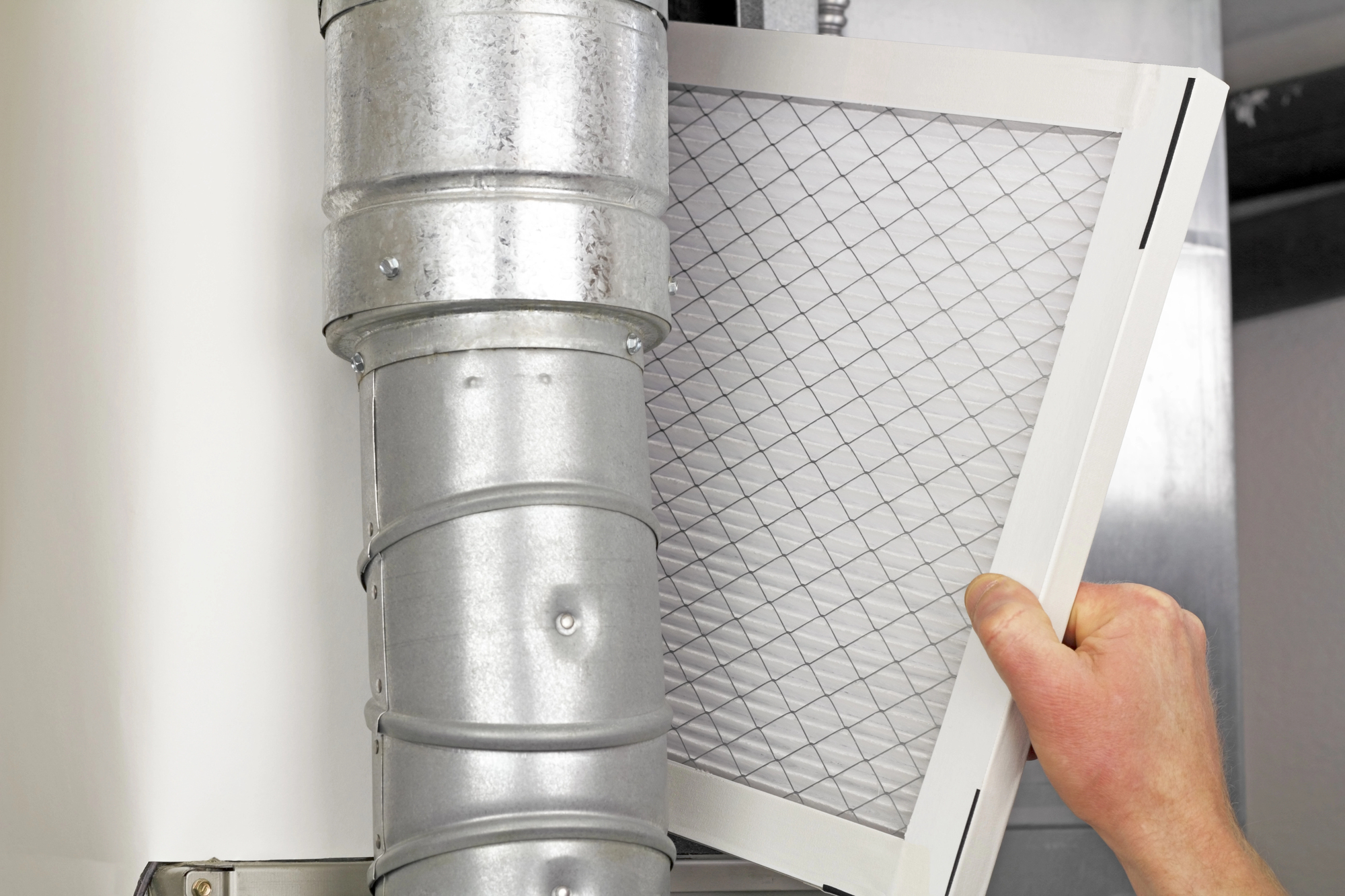 A hand removing a rectangular air filter from a duct, showing a metal duct with a flexed elbow joint in the background.