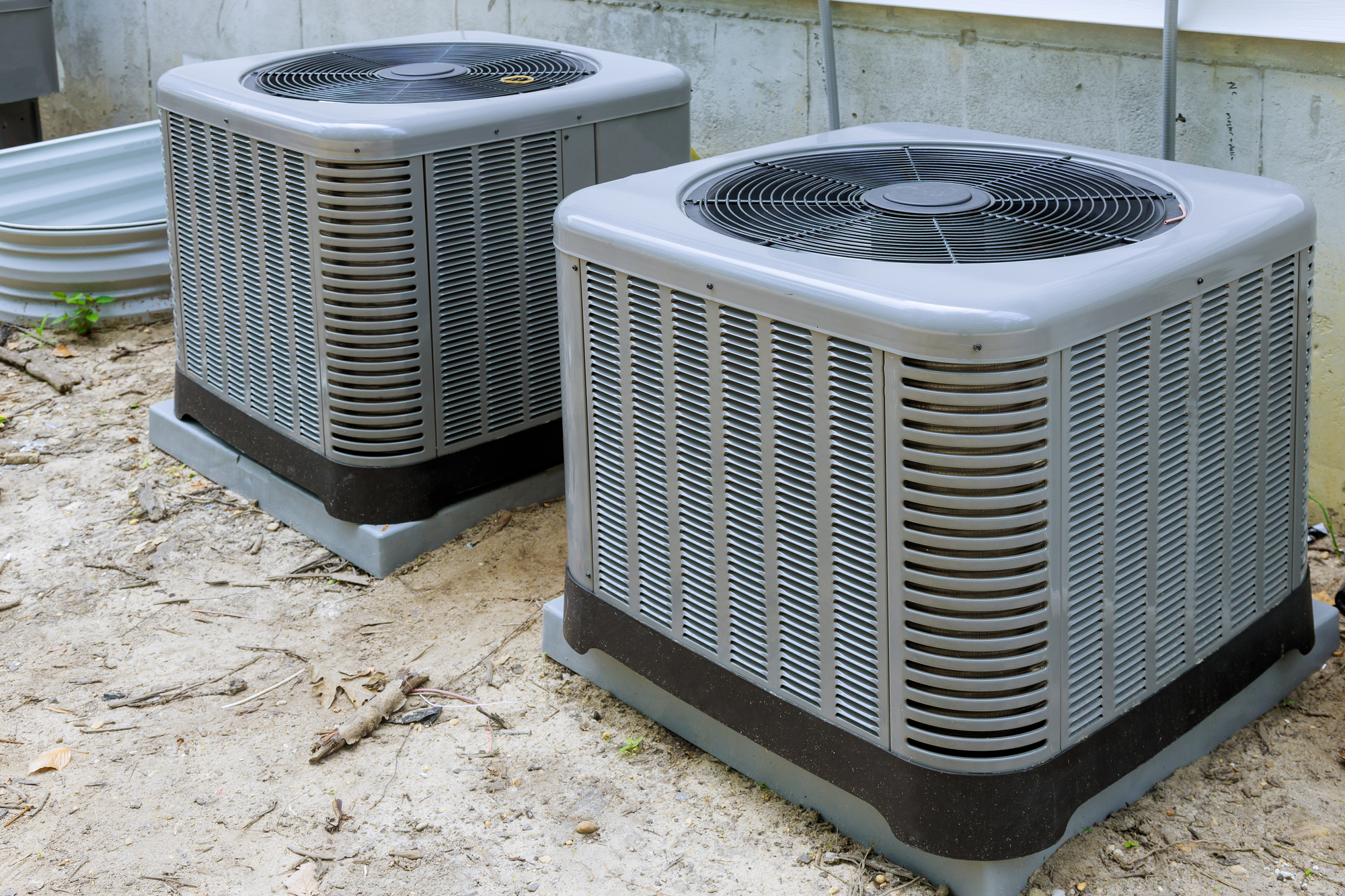 Two large outdoor air conditioning units sit side by side on a concrete base, surrounded by dirt and minimal vegetation.